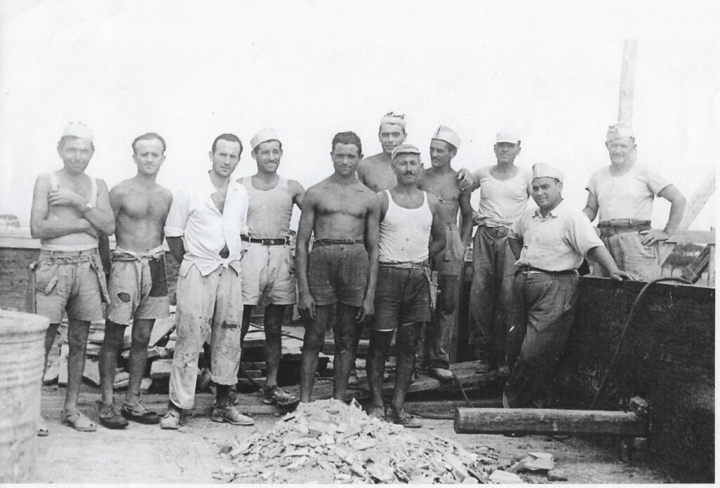 11 operai della Ditta Belotti posano a torso nudo o canottiera con i classici cappelli da muratore, tra tavole e attrezzi da lavoro